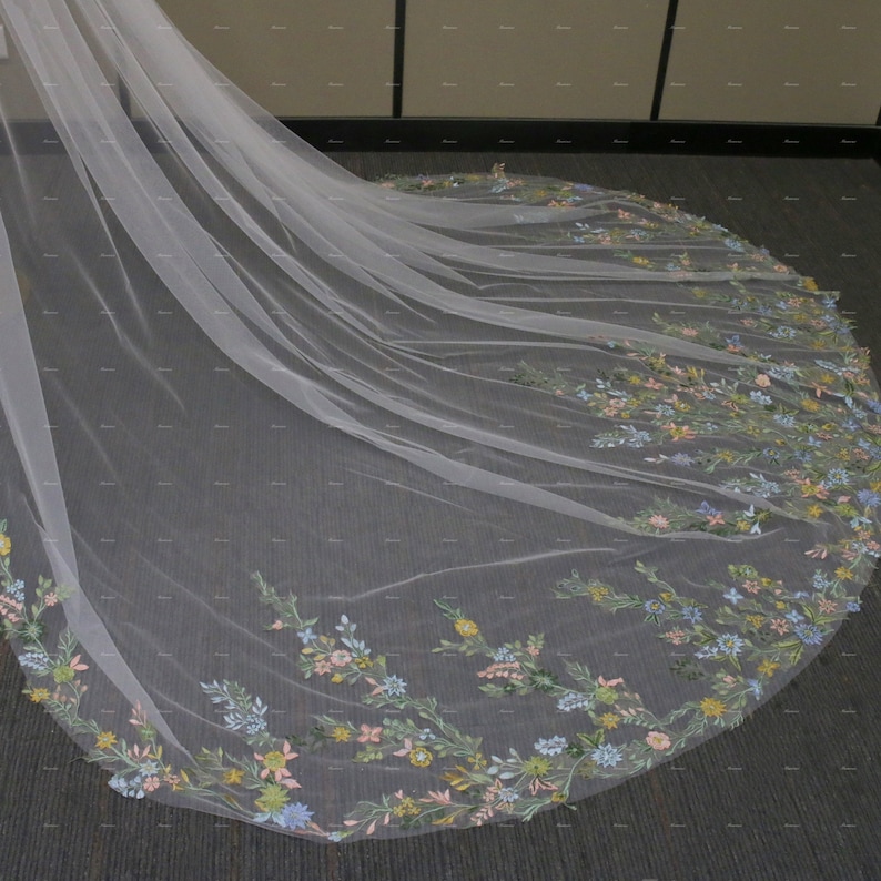 May include: A white tulle wedding veil with a floral embroidery design. The embroidery features pastel blue, pink, yellow, and green flowers.