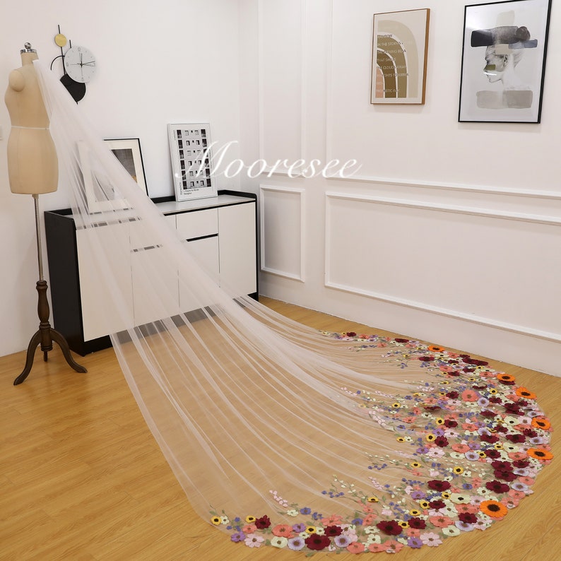 May include: A white tulle wedding veil with a floral design. The veil has a long train with a border of colorful embroidered flowers.