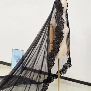 May include: A black bridal veil with intricate lace detailing. The veil is displayed on a mannequin, showcasing its flowing design and delicate floral lace trim along the edges. The veil is made of sheer fabric.
