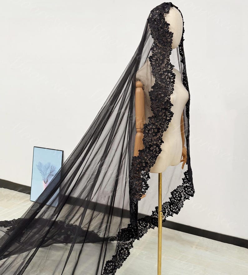 May include: A black bridal veil with intricate lace detailing. The veil is displayed on a mannequin, showcasing its flowing design and delicate floral lace trim along the edges. The veil is made of sheer fabric.