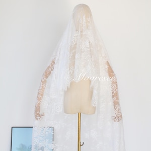 May include: A white lace wedding veil draped over a mannequin. The veil features intricate floral patterns and a delicate, scalloped edge. The word "Mooreseco" is visible on the veil. The veil is displayed on a gold stand against a white background.