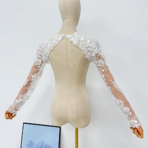 May include: A white, long-sleeved bridal bolero with intricate floral lace detailing. The bolero is displayed on a mannequin, showcasing its delicate design and sheer sleeves. The lace extends from the shoulders to the wrists, creating an elegant, romantic look.