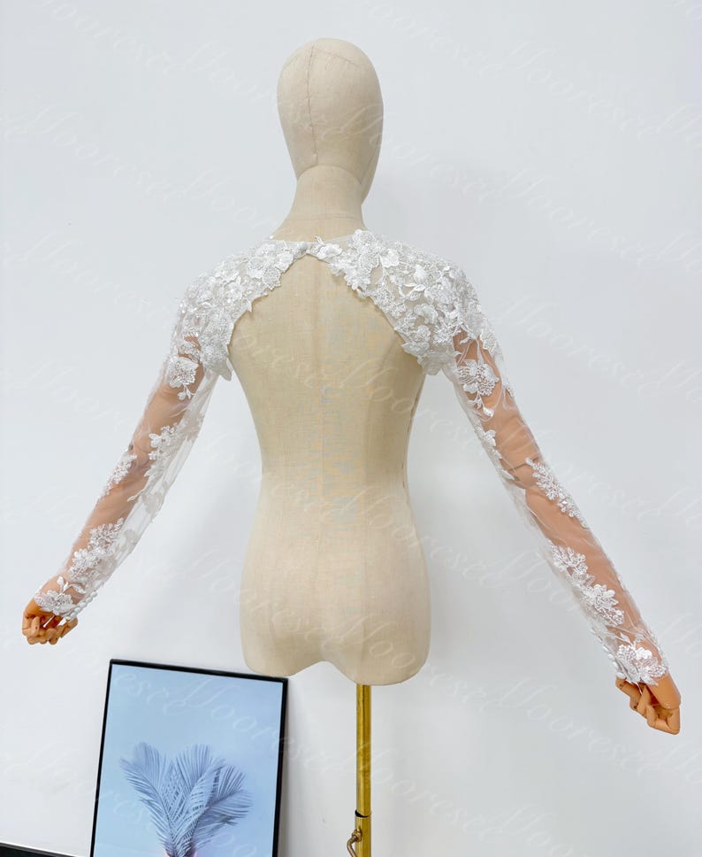 May include: A white, long-sleeved bridal bolero with intricate floral lace detailing. The bolero is displayed on a mannequin, showcasing its delicate design and sheer sleeves. The lace extends from the shoulders to the wrists, creating an elegant, romantic look.
