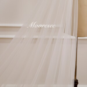 May include: White tulle fabric with a soft, flowing texture. The fabric is draped over a white surface and has a subtle sheen.