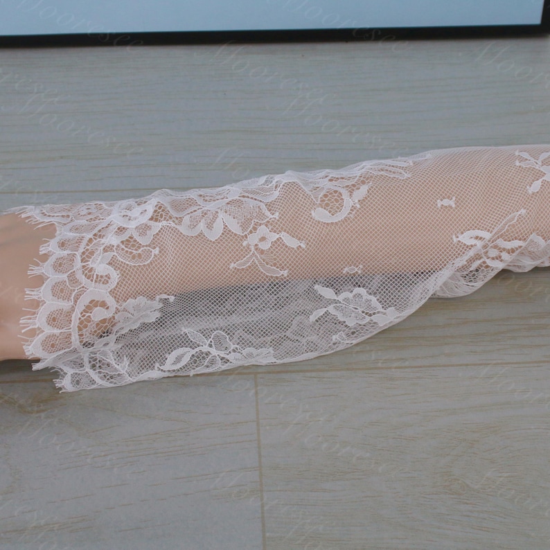 May include: Close-up of white lace fabric featuring floral patterns. The delicate lace has intricate designs and a scalloped edge. The fabric is semi-sheer, revealing the skin beneath. Suitable for bridal wear or craft projects.