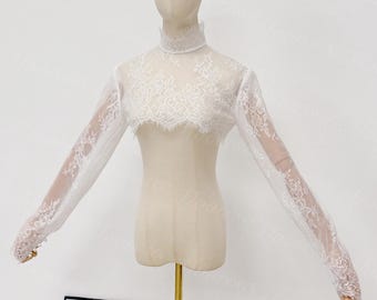 May include: A white lace bolero with long sleeves and a high, rounded collar. The delicate lace design covers the shoulders and sleeves, creating a sheer effect. The bolero is displayed on a mannequin.