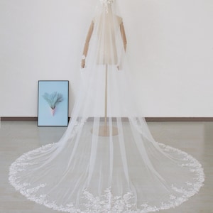 May include: A long, white bridal veil with floral lace detailing along the hem and a matching headpiece. The veil is displayed on a mannequin, showcasing its length and delicate design. The veil's train extends across the floor.