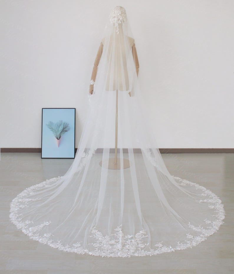 May include: A long, white bridal veil with floral lace detailing along the hem and a matching headpiece. The veil is displayed on a mannequin, showcasing its length and delicate design. The veil's train extends across the floor.