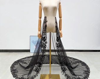 May include: A black tulle overskirt with floral lace detailing, displayed on a mannequin. The overskirt features a long train and intricate floral patterns along the edges and down the sides, creating an elegant and dramatic look.