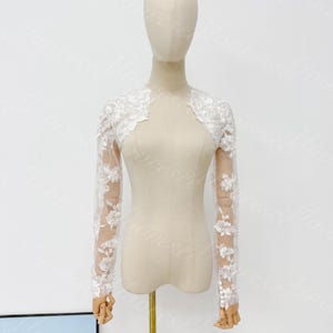 May include: A white, long-sleeved bolero jacket with floral lace detailing. The sheer sleeves feature delicate floral embroidery, and the bolero is displayed on a mannequin. The jacket is designed for formal occasions.