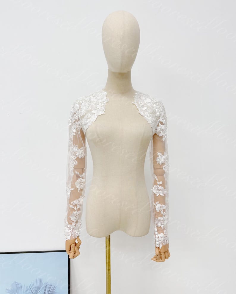 May include: A white, long-sleeved bolero jacket with floral lace detailing. The sheer sleeves feature delicate floral embroidery, and the bolero is displayed on a mannequin. The jacket is designed for formal occasions.