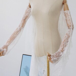 May include: Elegant white lace bridal gloves with floral detailing. The gloves are long, extending up the arms, and crafted from a delicate, sheer material. The gloves are displayed on a mannequin, highlighting their intricate design and graceful style.