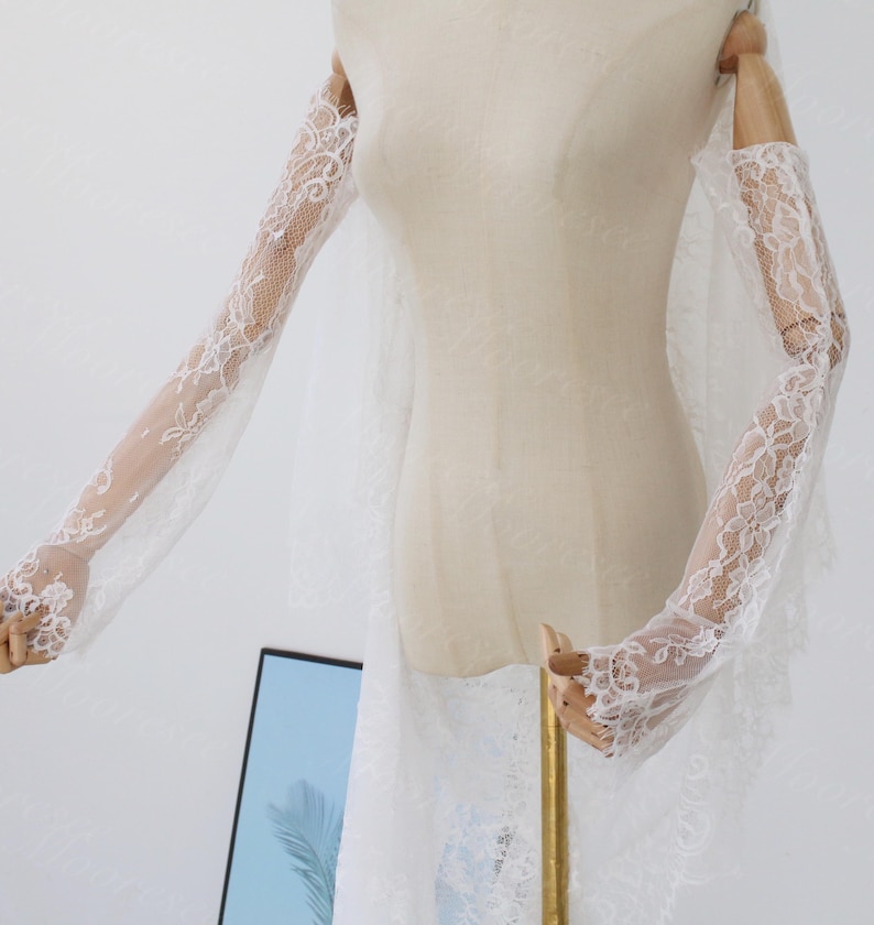 May include: Elegant white lace bridal gloves with floral detailing. The gloves are long, extending up the arms, and crafted from a delicate, sheer material. The gloves are displayed on a mannequin, highlighting their intricate design and graceful style.