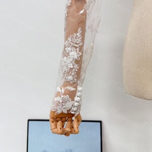 May include: A sheer white bridal sleeve with intricate floral lace and sequin detailing. The sleeve features a row of white buttons at the wrist and is displayed on a wooden hand mannequin. The design is elegant and suitable for weddings.