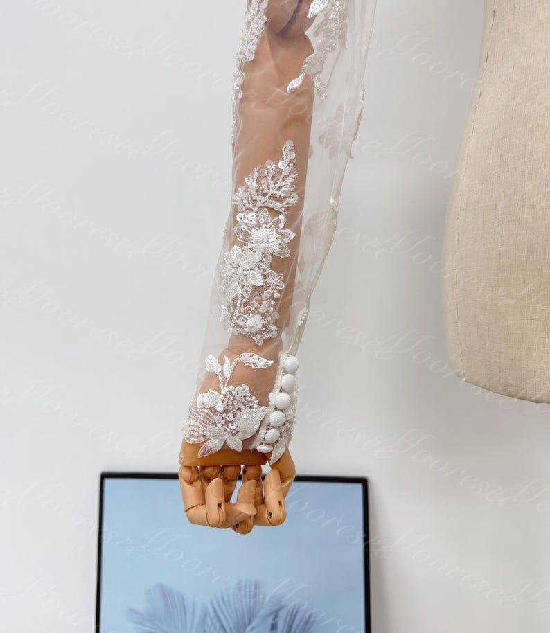 May include: A sheer white bridal sleeve with intricate floral lace and sequin detailing. The sleeve features a row of white buttons at the wrist and is displayed on a wooden hand mannequin. The design is elegant and suitable for weddings.