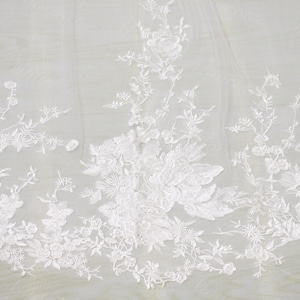 以下が含まれることがあります： 白い花のレース生地で、刺繍された花と葉が特徴です。繊細なデザインは透明な背景に配置され、質感とエレガントな外観を作り出しています。ブライダルウェアや装飾プロジェクトに最適です。