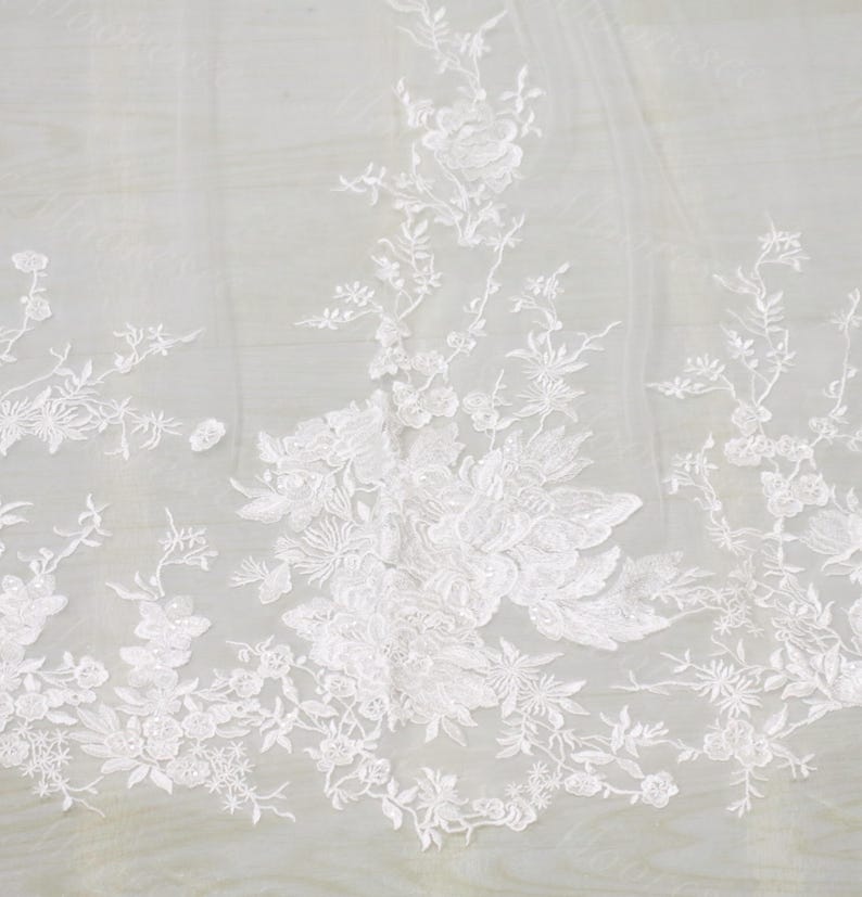 以下が含まれることがあります： 白い花のレース生地で、刺繍された花と葉が特徴です。繊細なデザインは透明な背景に配置され、質感とエレガントな外観を作り出しています。ブライダルウェアや装飾プロジェクトに最適です。