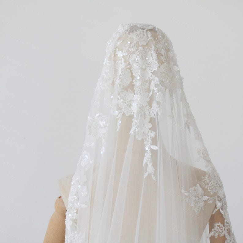 May include: A white bridal veil with floral lace and sequin embellishments. The veil is draped over a mannequin head, showcasing the intricate details of the lace design. The veil is made of a sheer, lightweight fabric.