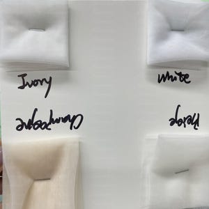 May include: A color swatch card displaying samples of tulle fabric in ivory, champagne, white, and beige. Each sample is pinned to the card. Handwritten labels identify each color. The background is white.