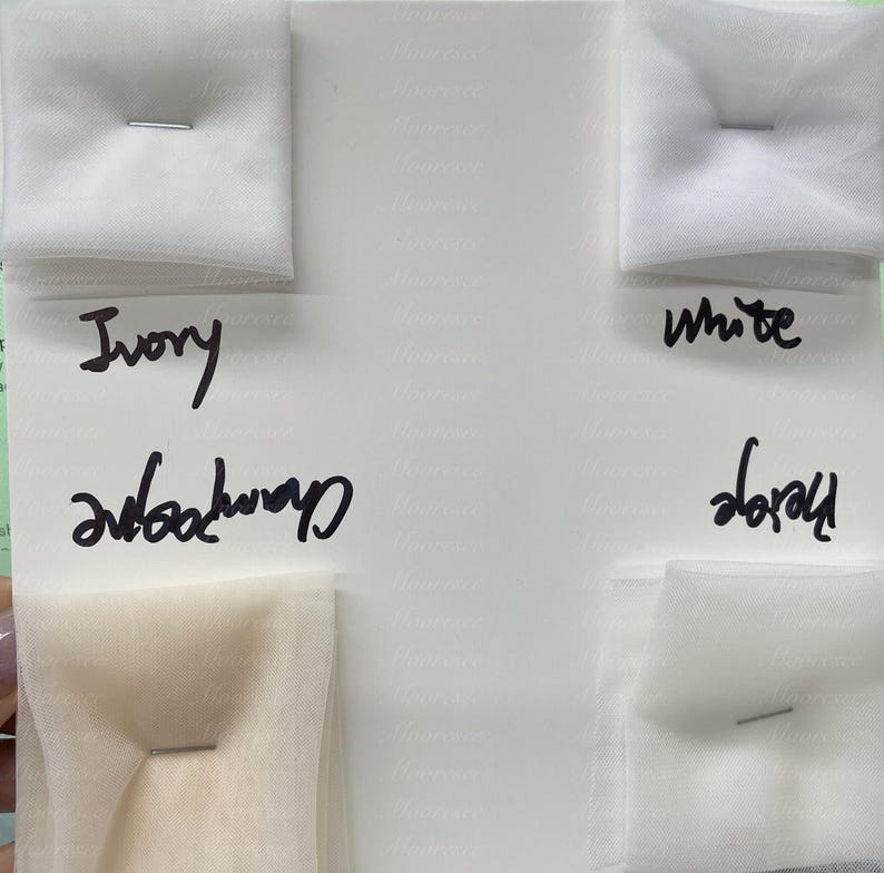 May include: A color swatch card displaying samples of tulle fabric in ivory, champagne, white, and beige. Each sample is pinned to the card. Handwritten labels identify each color. The background is white.
