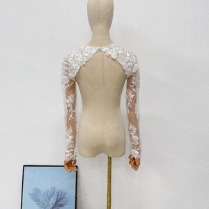 May include: A white lace bridal bolero with long sleeves. The bolero features intricate floral lace detailing on the sleeves and shoulders. The back has a keyhole design. The bolero is displayed on a mannequin against a white background.