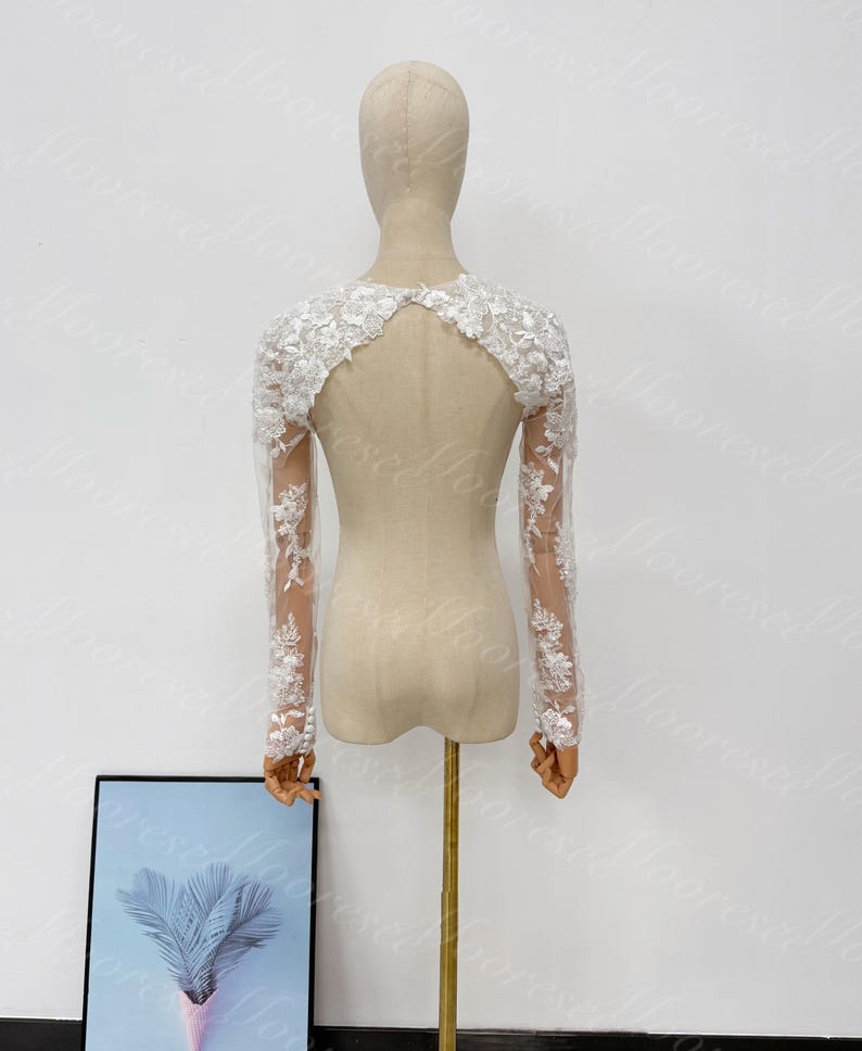 May include: A white lace bridal bolero with long sleeves. The bolero features intricate floral lace detailing on the sleeves and shoulders. The back has a keyhole design. The bolero is displayed on a mannequin against a white background.