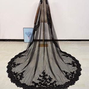 May include: A long, black bridal veil with intricate floral lace detailing along the edges. The veil is displayed on a mannequin, showcasing its full length and delicate design. The lace adds a touch of elegance.