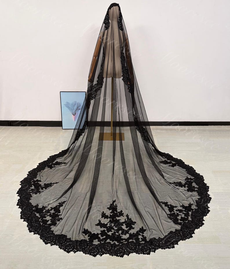 May include: A long, black bridal veil with intricate floral lace detailing along the edges. The veil is displayed on a mannequin, showcasing its full length and delicate design. The lace adds a touch of elegance.