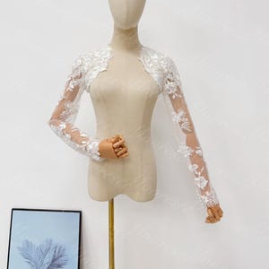 May include: A white lace bolero with long sleeves. The bolero features delicate floral embroidery and a sheer design. The garment is displayed on a mannequin, showcasing its elegant and intricate details. Perfect for weddings or special occasions.