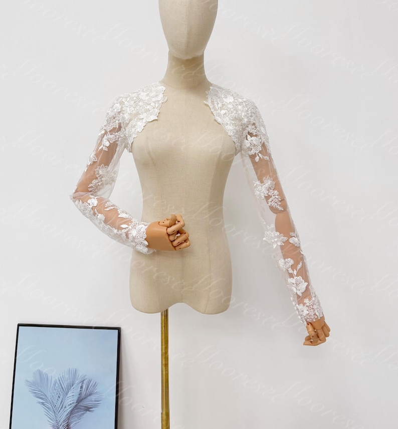 May include: A white lace bolero with long sleeves. The bolero features delicate floral embroidery and a sheer design. The garment is displayed on a mannequin, showcasing its elegant and intricate details. Perfect for weddings or special occasions.