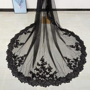 May include: A long, black bridal veil with intricate floral lace detailing along the edges and scattered across the sheer fabric. The veil is spread out, showcasing its full length and design.