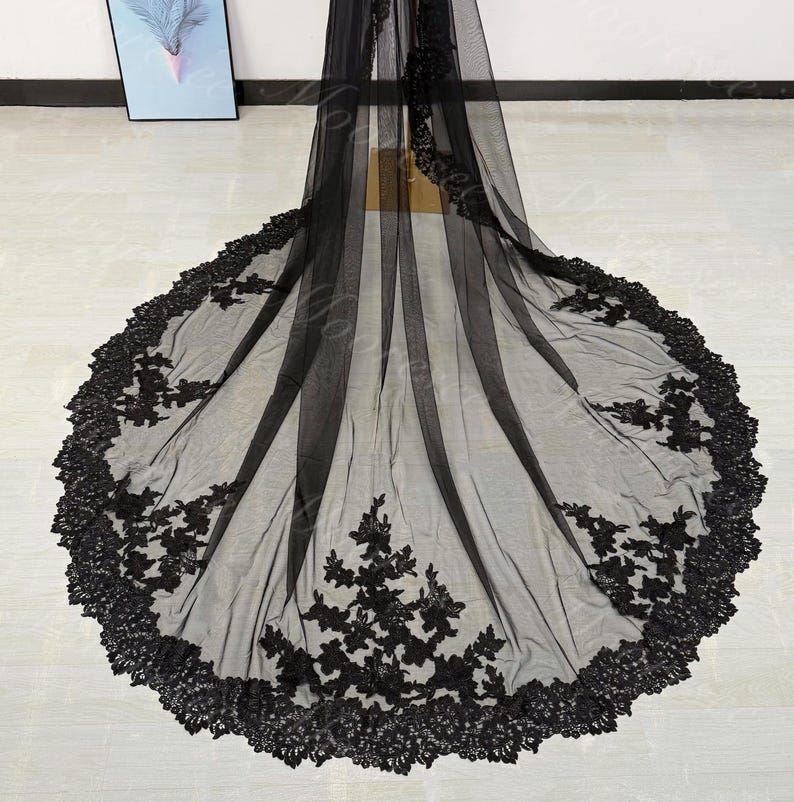 May include: A long, black bridal veil with intricate floral lace detailing along the edges and scattered across the sheer fabric. The veil is spread out, showcasing its full length and design.