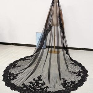 May include: A long, black bridal veil with intricate floral lace detailing along the edges. The veil is draped over a mannequin, showcasing its flowing design and delicate texture. The veil's length extends to the floor, creating a dramatic effect.