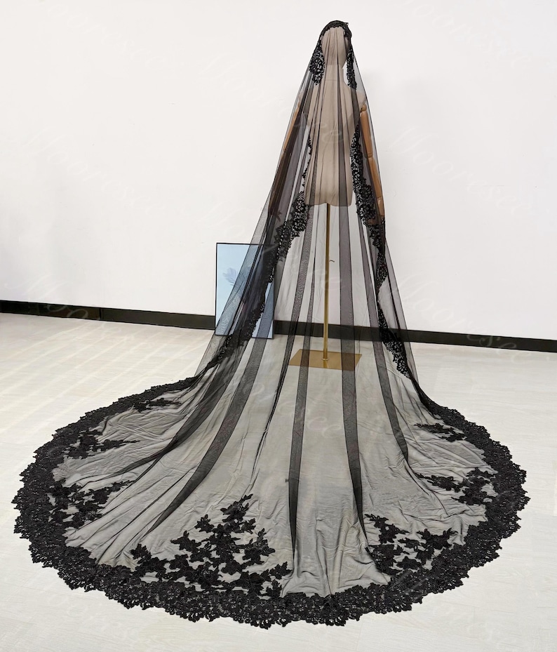 May include: A long, black bridal veil with intricate floral lace detailing along the edges. The veil is draped over a mannequin, showcasing its flowing design and delicate texture. The veil's length extends to the floor, creating a dramatic effect.