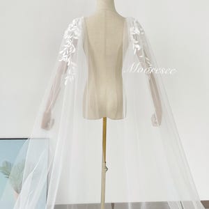 May include: A sheer white bridal cape with delicate floral embroidery on the shoulders. The cape is displayed on a mannequin, showcasing its flowing design. The word "Mooresee" is printed on the cape.