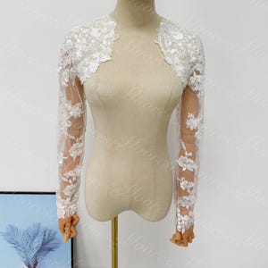 May include: A white lace bolero jacket with long sleeves. The jacket features floral lace detailing on the shoulders and sleeves. The sleeves are made of a sheer fabric, and the jacket is displayed on a mannequin.