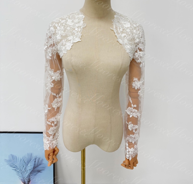 May include: A white lace bolero jacket with long sleeves. The jacket features floral lace detailing on the shoulders and sleeves. The sleeves are made of a sheer fabric, and the jacket is displayed on a mannequin.