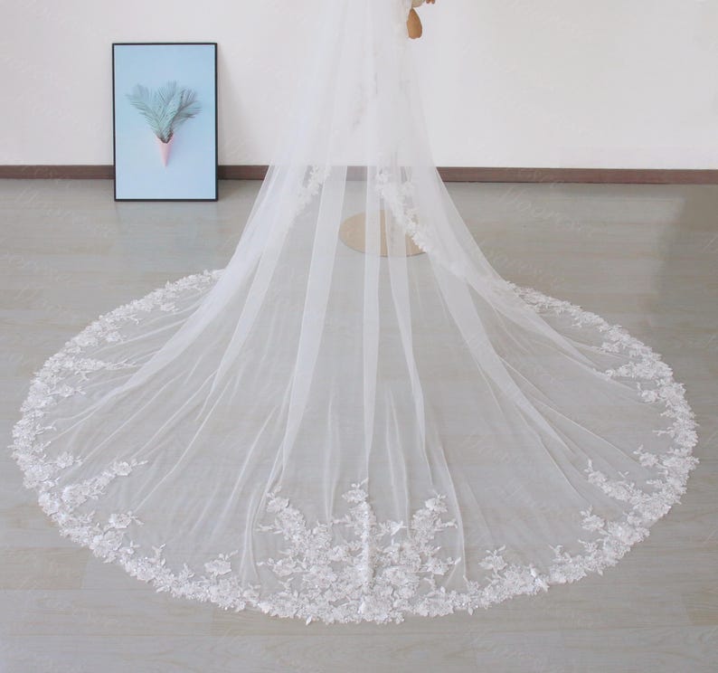 May include: A long, white bridal veil with a floral lace trim. The veil is made of a sheer fabric and drapes to the floor. The lace trim features delicate, three-dimensional floral designs.
