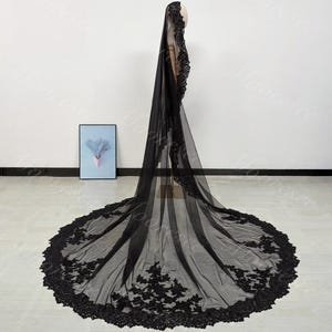 May include: A long, black bridal veil with intricate floral lace detailing along the edges. The veil is made of a sheer fabric and is displayed on a mannequin, showcasing its full length and design.