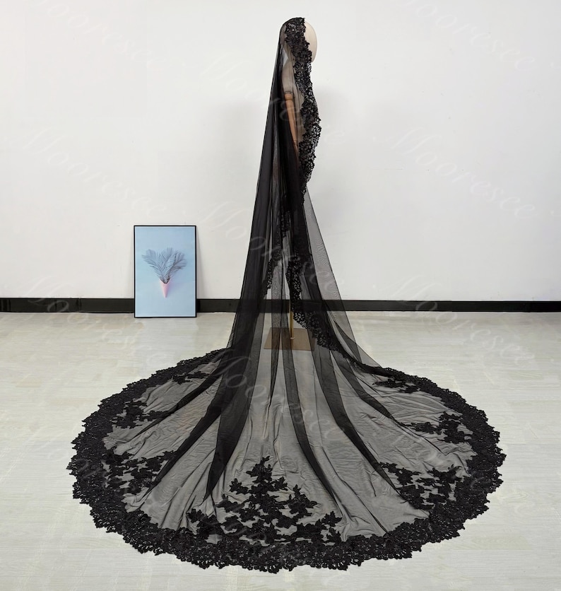 May include: A long, black bridal veil with intricate floral lace detailing along the edges. The veil is made of a sheer fabric and is displayed on a mannequin, showcasing its full length and design.
