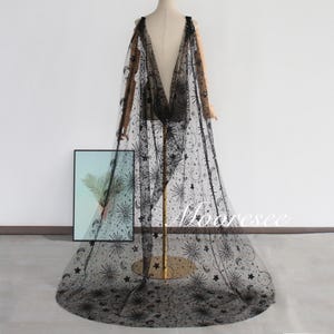 May include: A black, sheer cape with a celestial pattern of stars, moons, and suns. The cape is draped over a mannequin, showcasing its flowing design. The cape is floor-length and features a detailed, all-over print.