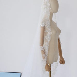 May include: A white bridal veil with floral lace and sequin embellishments. The veil is long and sheer, with delicate detailing along the edges and sleeves. The veil is displayed on a mannequin.