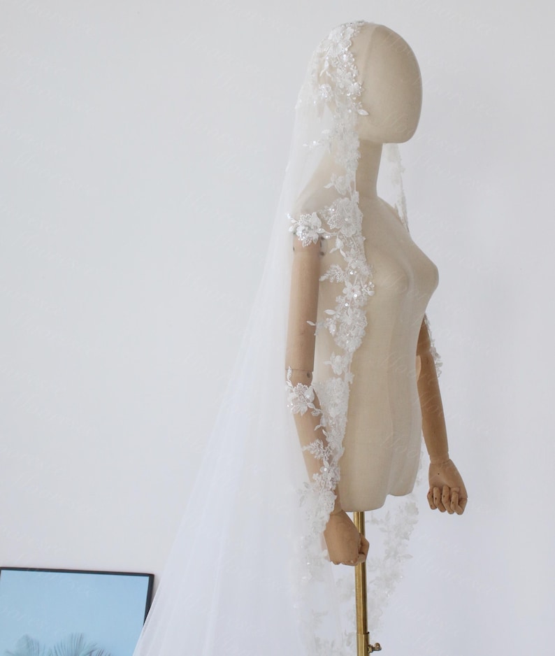 May include: A white bridal veil with floral lace and sequin embellishments. The veil is long and sheer, with delicate detailing along the edges and sleeves. The veil is displayed on a mannequin.
