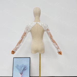May include: A white lace bridal bolero with long sleeves, displayed on a mannequin. The bolero features floral lace detailing on the sleeves and shoulders, with a sheer back. The mannequin is on a gold stand, with a framed picture in the foreground.