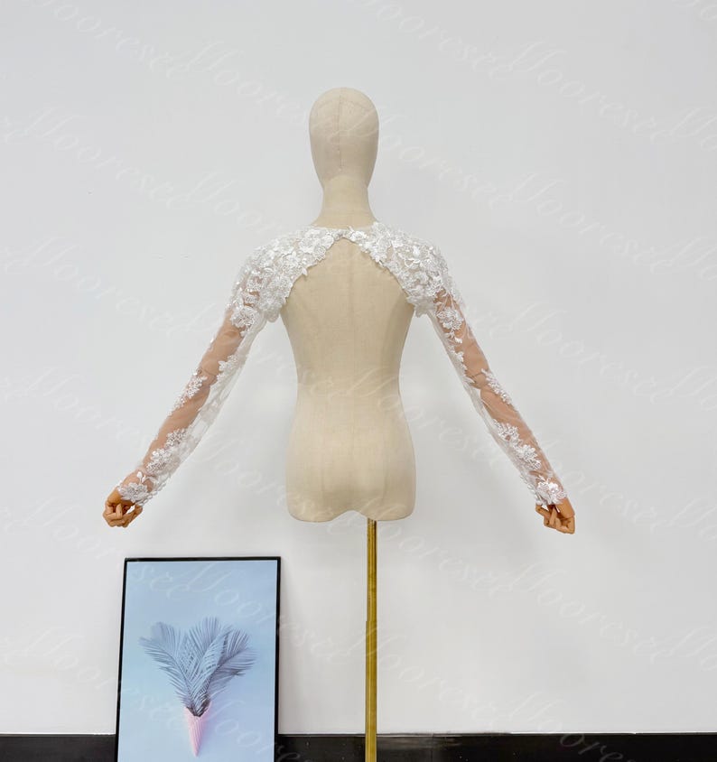 May include: A white lace bridal bolero with long sleeves, displayed on a mannequin. The bolero features floral lace detailing on the sleeves and shoulders, with a sheer back. The mannequin is on a gold stand, with a framed picture in the foreground.