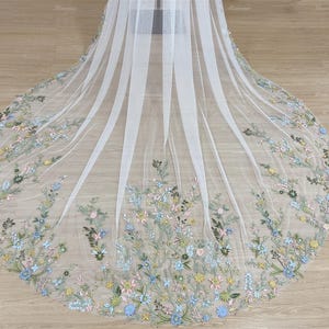 May include: A white tulle veil with delicate floral embroidery in shades of blue, green, and pink. The veil is gathered at the top and falls in soft layers.