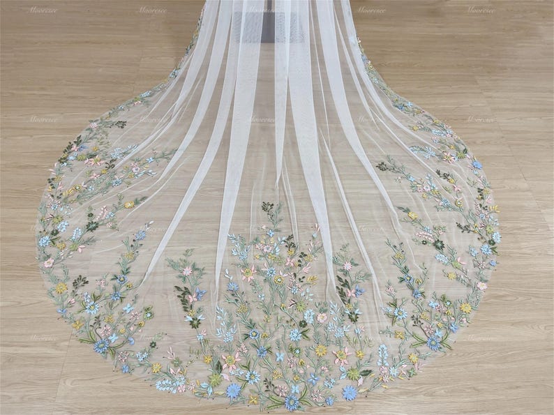 May include: A white tulle veil with delicate floral embroidery in shades of blue, green, and pink. The veil is gathered at the top and falls in soft layers.