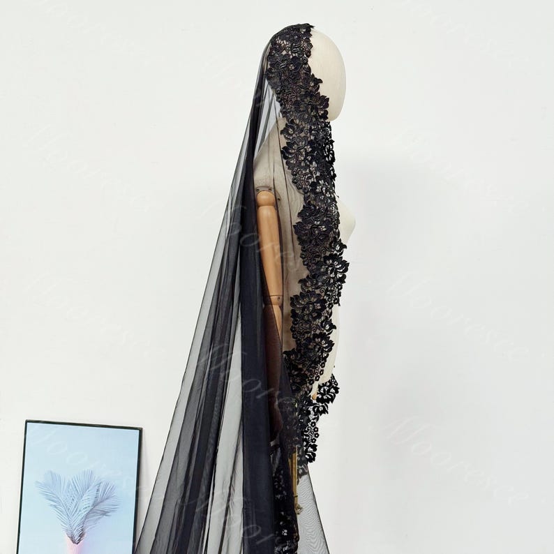 May include: A black bridal veil with intricate floral lace detailing along the edge. The veil is long and flowing, made of a sheer fabric. The lace is a darker shade of black, creating a contrast against the sheer material.