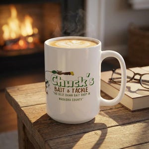 May include: White ceramic mug with a handle, filled with a latte. The mug features the text "Chuck's Bait & Tackle" in green and black, with a graphic of a fishing lure. The mug is on a wooden surface.