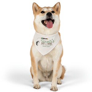 Könnte beinhalten: Ein tanfarbener und weißer Shiba Inu Hund, der ein weißes Bandana mit dem Text "Chuck's Bait & Tackle - The Best Damn Bait Shop in Wabash County" in grüner und brauner Schrift trägt. Der Hund sitzt mit offenem Maul und herausgestreckter Zunge.